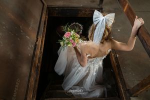 Bride in white lace gown holding pink floral bouquet descends wooden stairs, viewed from above, with bow veil.