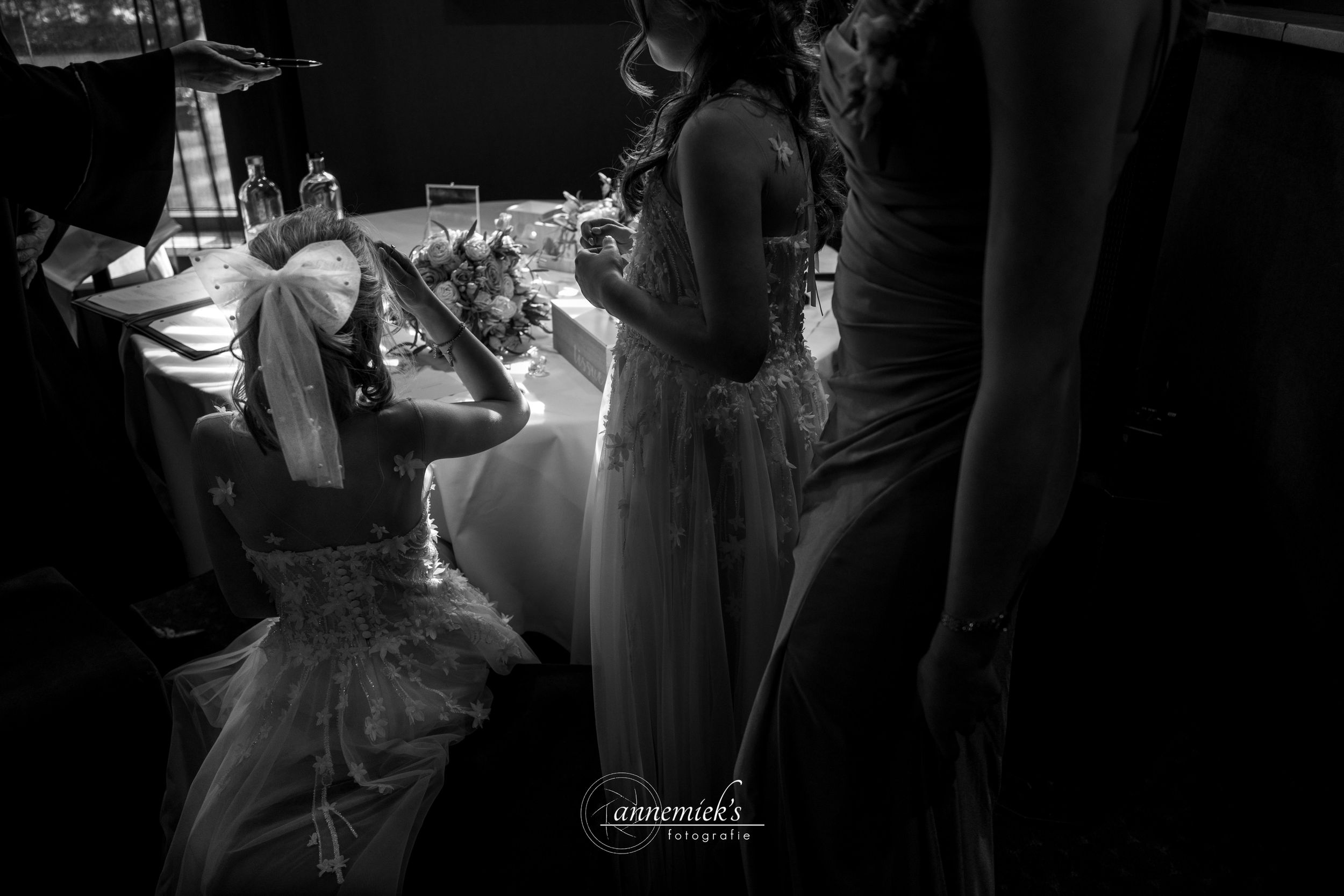 Black and white wedding photo of bride kneeling at reception table with guests in elegant gowns nearby.