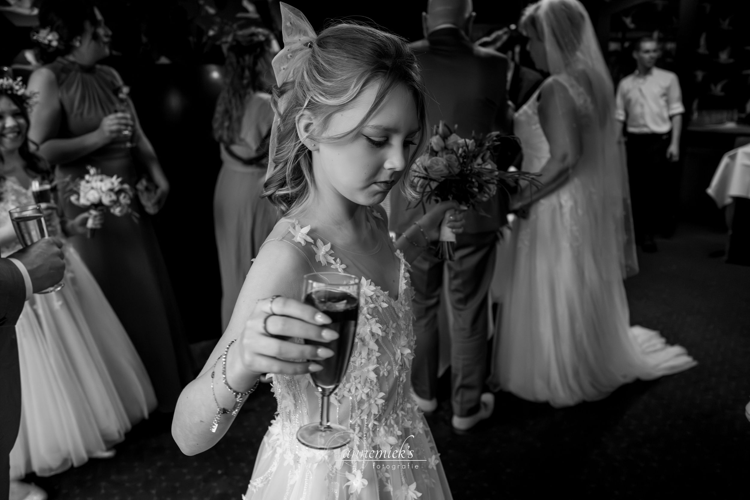A young flower girl in a white lace dress holds a wine glass at a wedding reception in black and white.