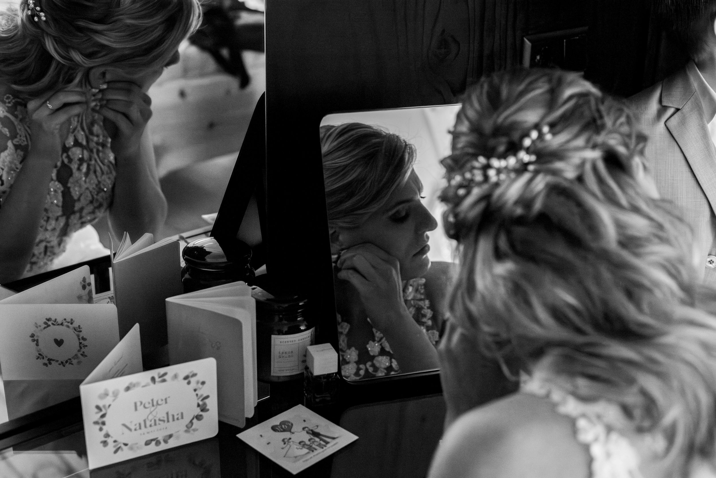 Black and white photo of a bride adjusting her earring in a mirror, with elegant updo hairstyle and floral hair accessory.