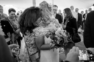 Black and white photo of a bride in a lace gown joyfully hugging a guest while holding a bouquet at a wedding reception.