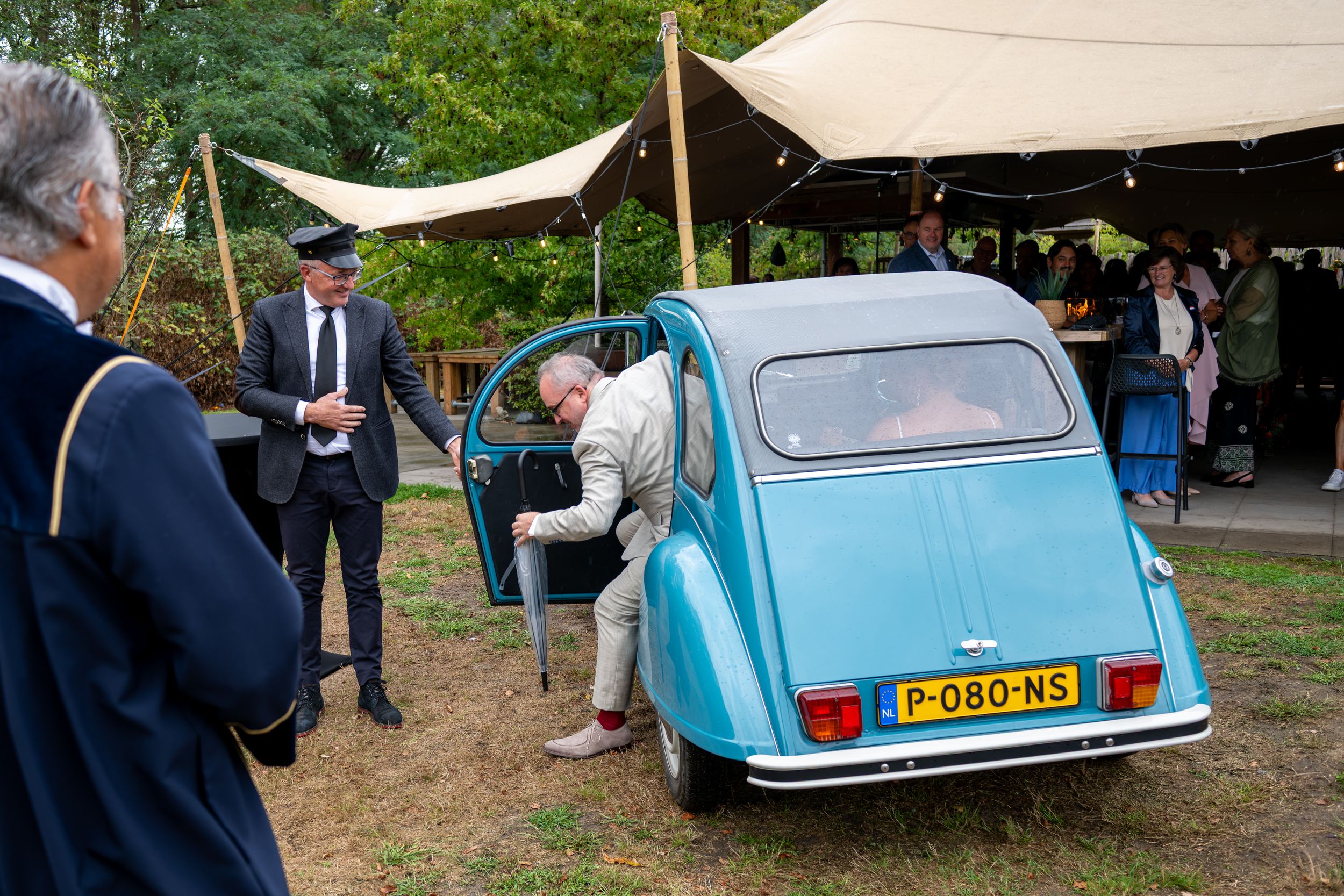 An elderly man exits a vintage blue Citroën 2CV at an outdoor event while suited guests look on.