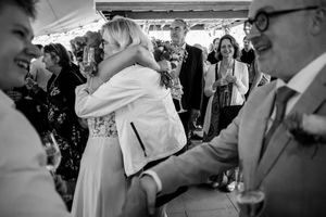 Black and white wedding photo of bride embracing guest at reception while guests laugh and celebrate around them.