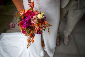 Bride holding vibrant wedding bouquet with purple orchids, orange, and pink flowers in colorful bridal arrangement.