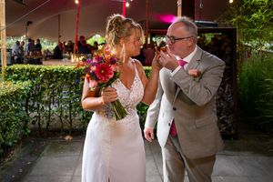 Groom kisses bride's hand outdoors at wedding reception, bride holds vibrant red and pink floral bouquet.