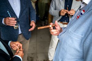 Men in blazers holding cigars at a formal outdoor gathering, one wearing a decorative lapel pin.