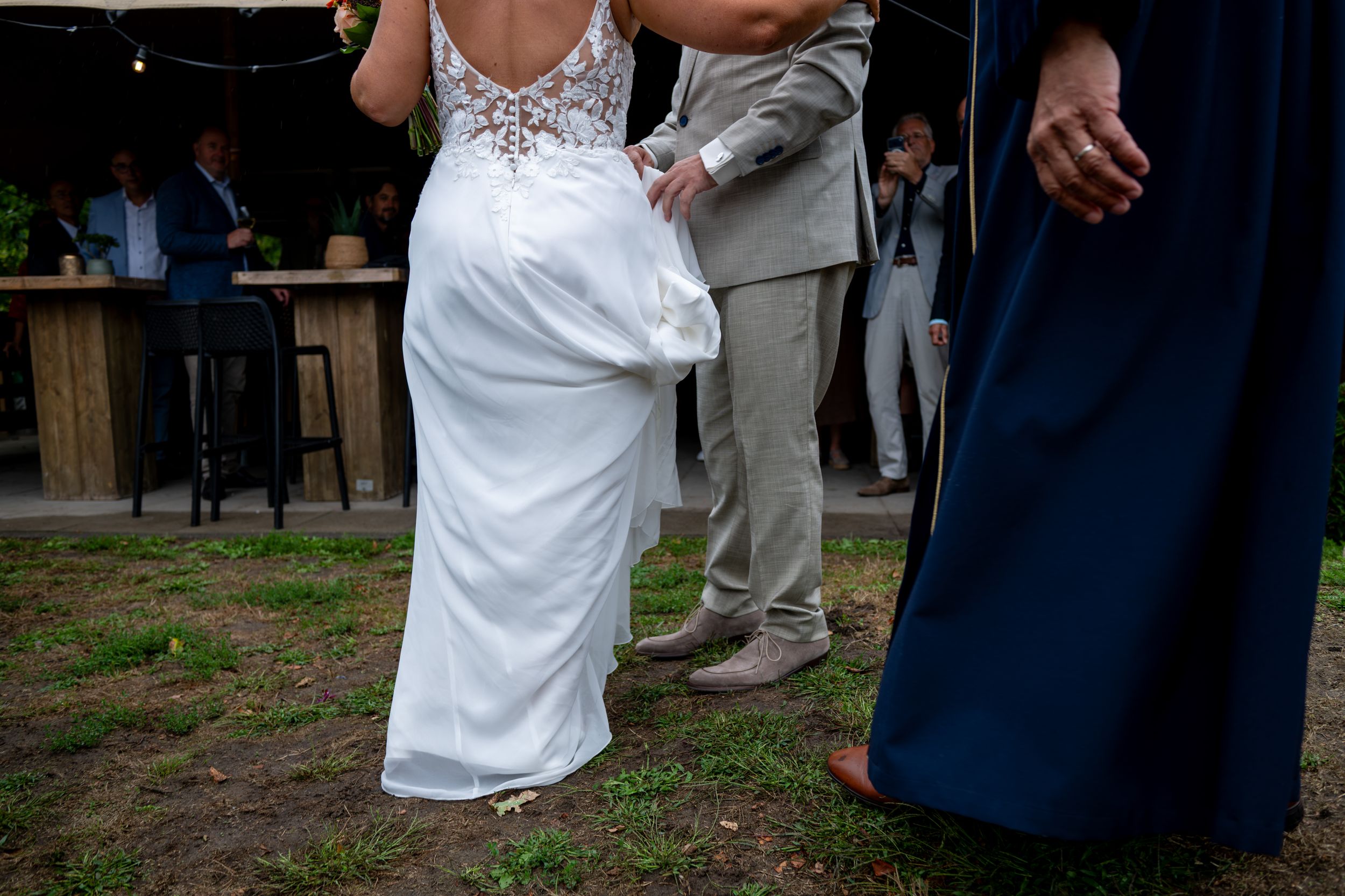 Bride in white lace gown stands barefoot on grass with groom in tan suit during outdoor wedding ceremony.