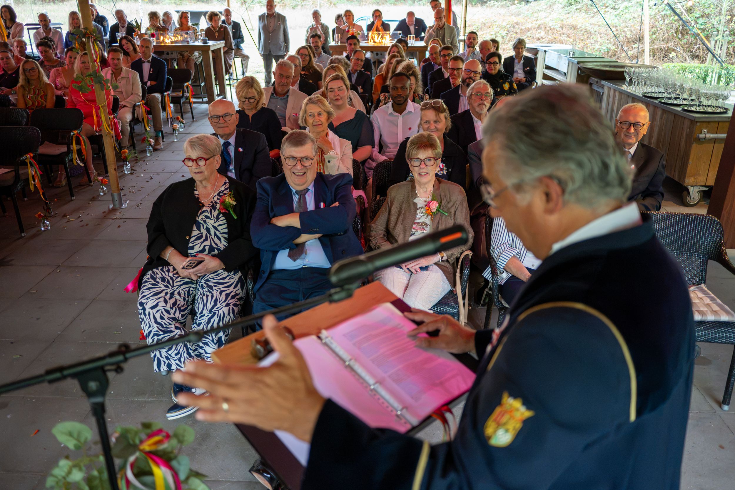 A speaker addresses a seated audience at an outdoor ceremony under a pavilion during a formal event.