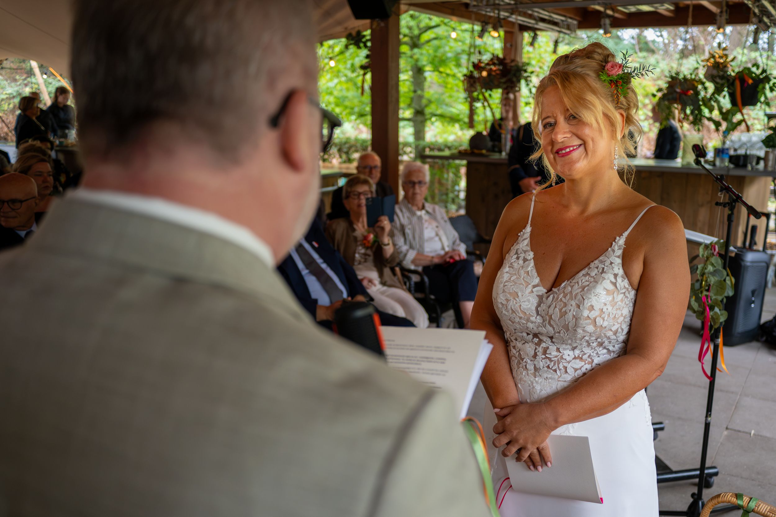 Bride smiling joyfully during outdoor wedding ceremony while exchanging vows with groom in tan suit.