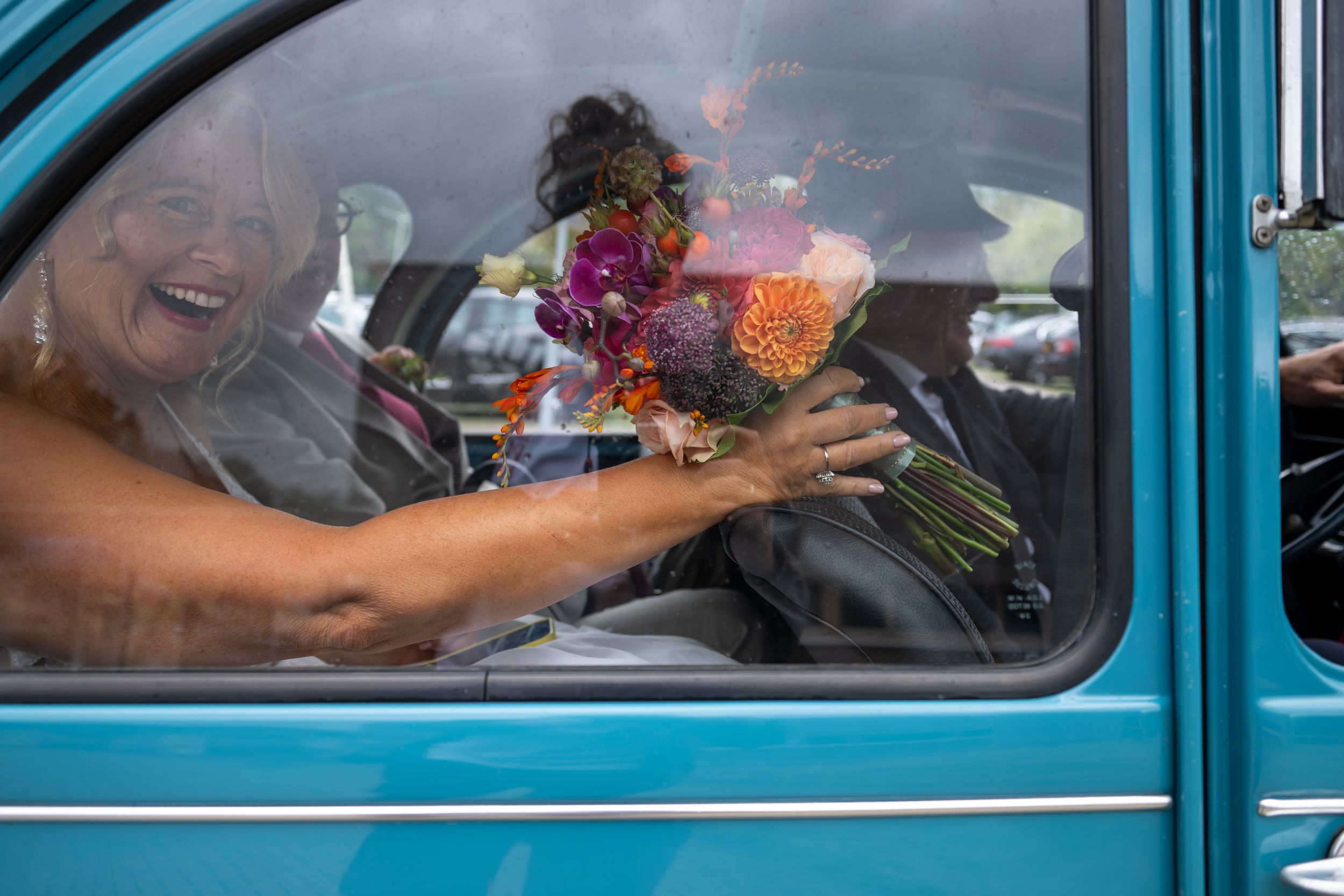 Woman laughing in vintage teal car holding colorful flower bouquet through window on sunny day.