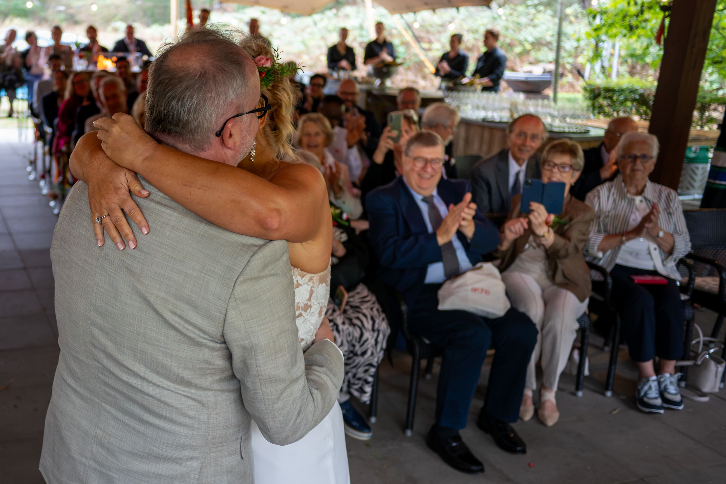 Bride and groom share a warm embrace during wedding ceremony while guests applaud and celebrate in outdoor venue.