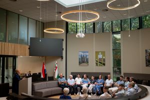 Modern atrium with circular lighting fixtures hosts a speaker addressing seated audience in a contemporary building.