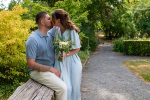 Couple sharing a romantic kiss in a lush garden, bride in light blue dress holding white bouquet, groom in casual attire.