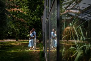 Couple embracing outdoors near a glass greenhouse surrounded by lush green trees, reflected in the greenhouse windows.
