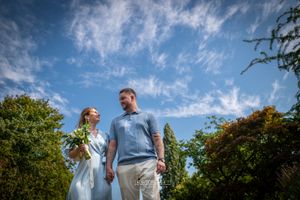 Couple holding hands and smiling outdoors under a bright blue sky with wispy clouds, surrounded by lush green trees.