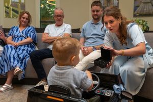 Woman in blue dress crouches to interact with toddler in toy car while family watches in modern lounge.