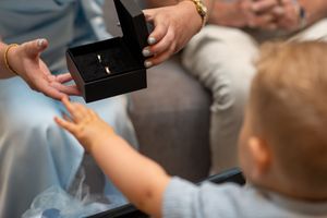 A toddler reaches for an open ring box held by an adult, revealing a gold wedding band inside.
