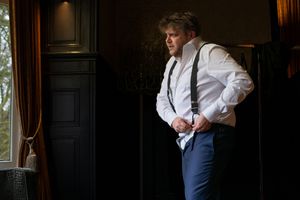 A person in formal attire stands in an elegant room with dark wood paneling and dramatic lighting.
