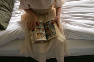Close-up view of someone sitting on white bedding while holding a book.