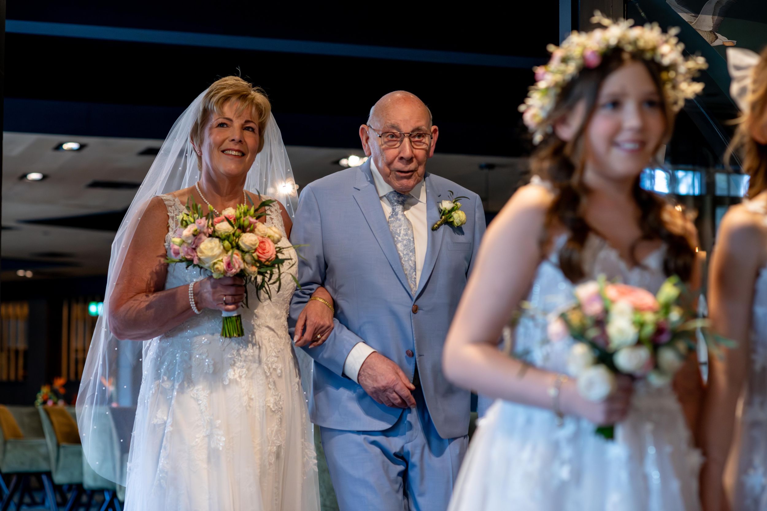 Bride in white gown walks down aisle with elderly man in blue suit, as flower girl leads the way at wedding ceremony.