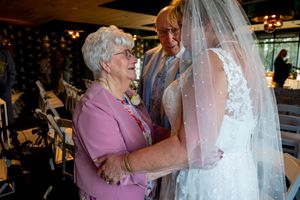 Elderly woman in pink adjusts bride's lace wedding dress and veil at indoor wedding venue.