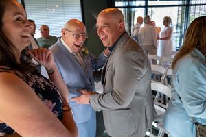 People laughing and shaking hands at a festive indoor gathering with guests in formal attire.