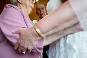 Elderly couple joyfully greeting a bride in a white wedding gown at a wedding reception celebration.