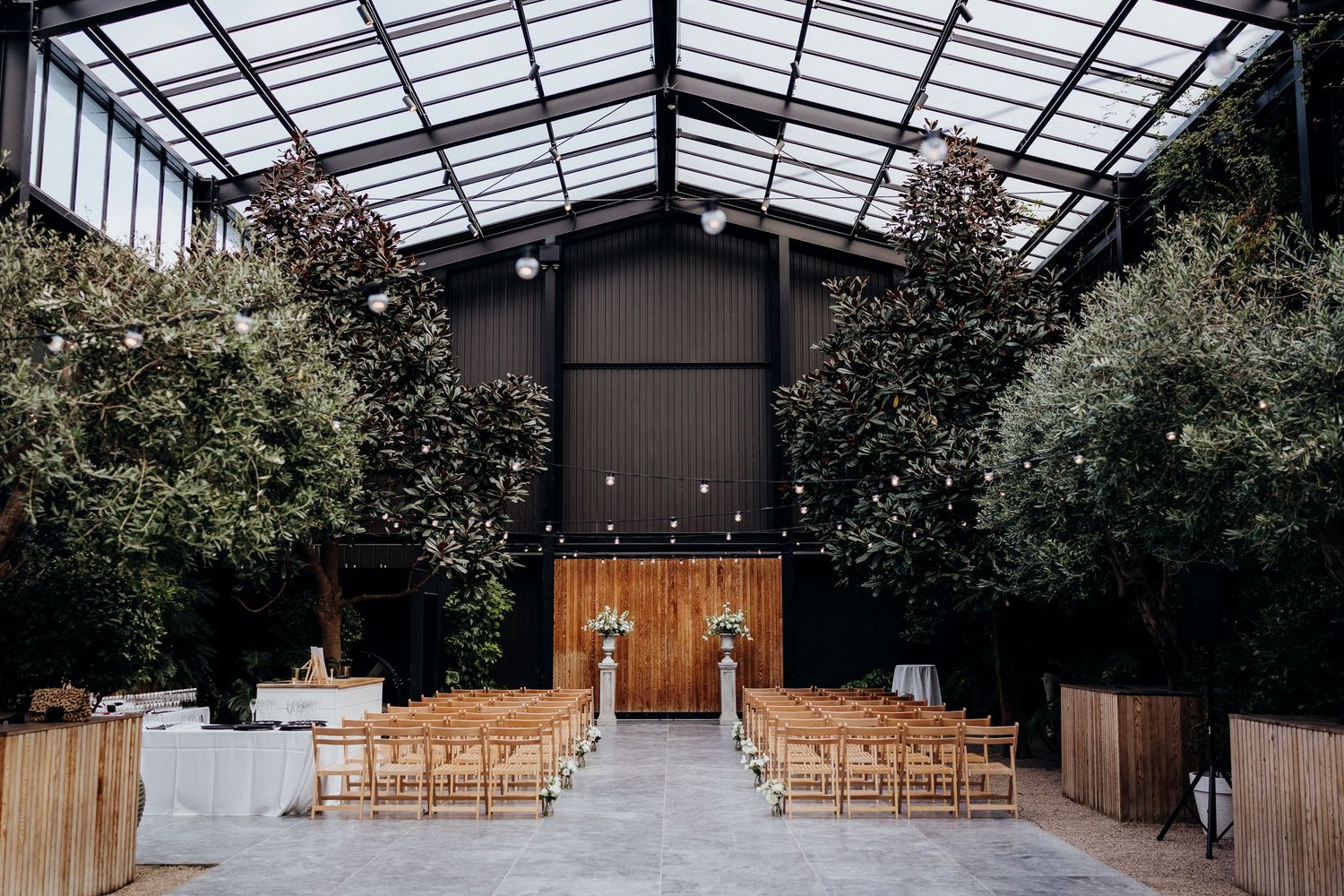 Interior view of a modern glass-roofed greenhouse space with wooden chairs set up in rows for a wedding ceremony.