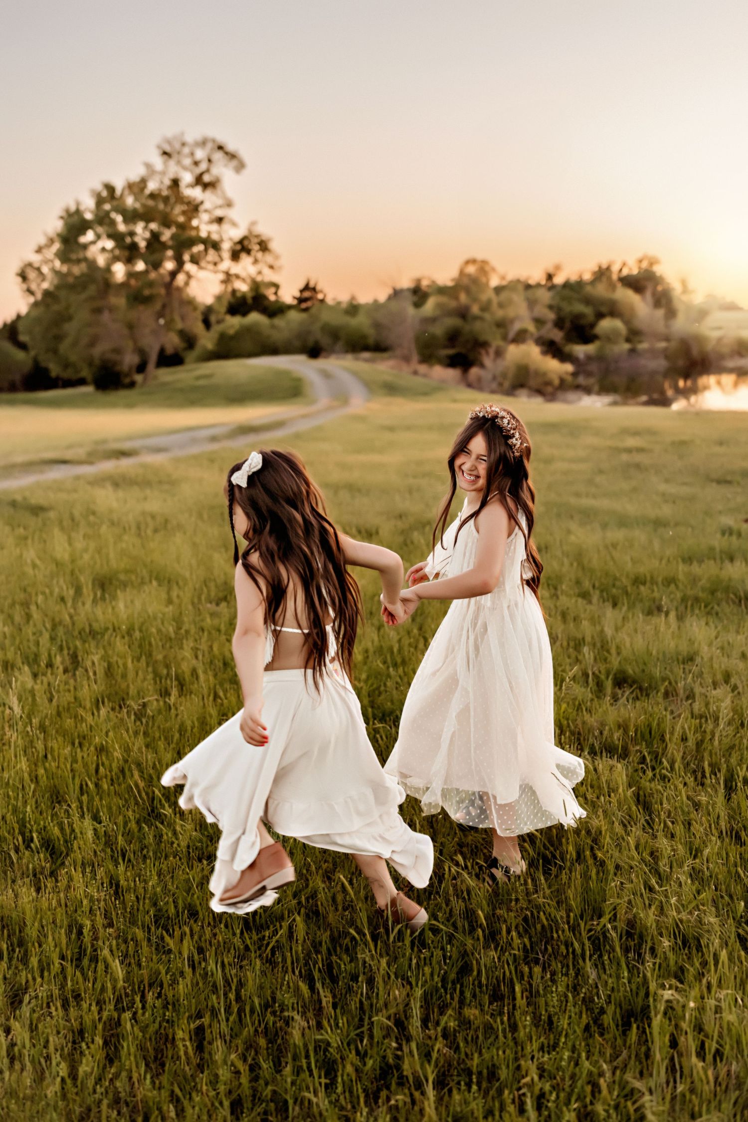 Two women in white dresses dancing together in a grassy field at sunset.