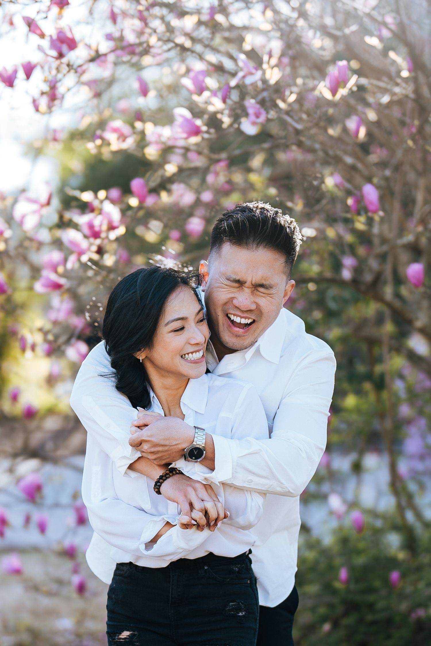 A joyful couple in white shirts share a candid laughing moment together under blooming pink magnolia blossoms.