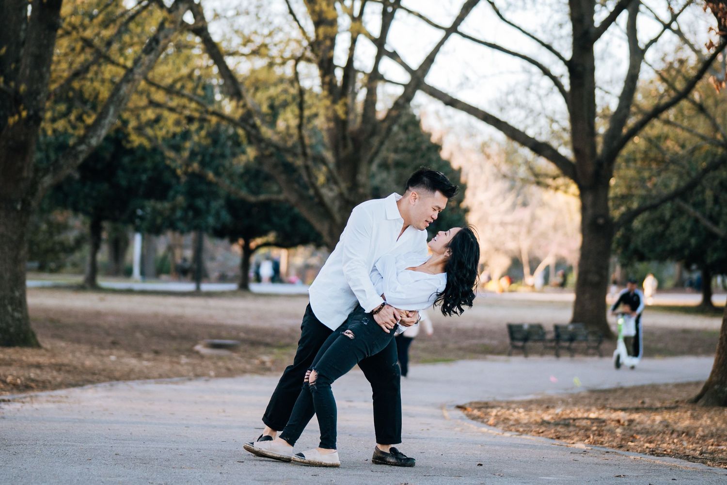 A couple in casual attire holds hands while walking through a park filled with fallen autumn leaves.
