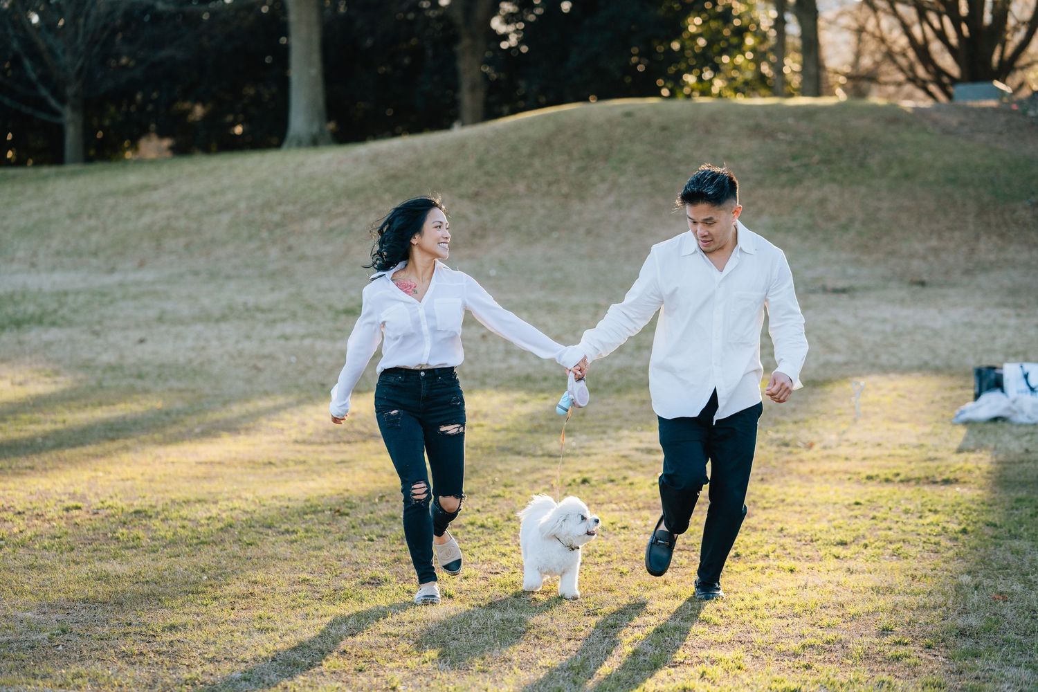 A couple walks hand in hand with their small white dog through a sunlit park.