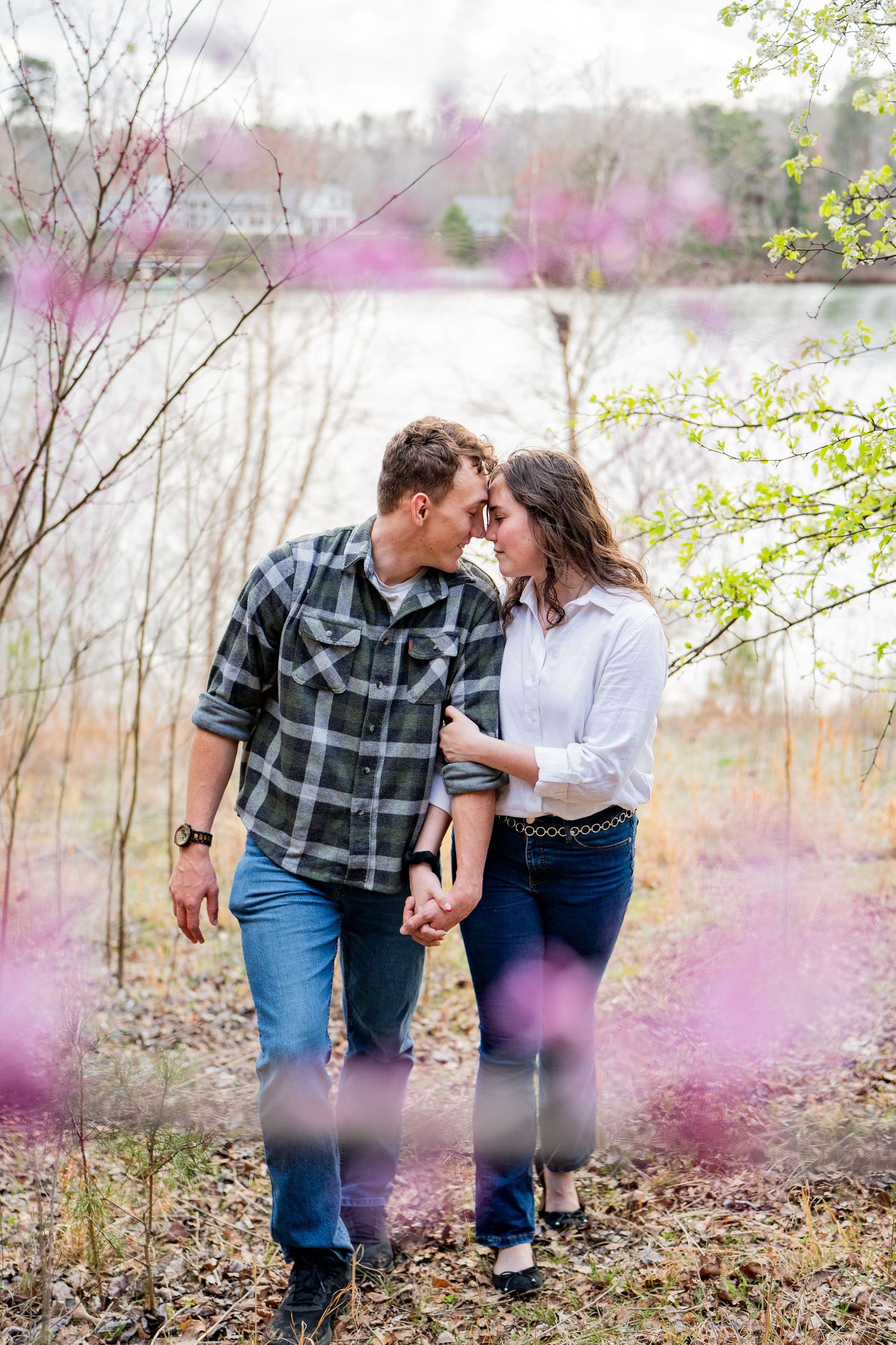 A couple shares an intimate moment in nature surrounded by soft pink cherry blossoms and spring foliage.
