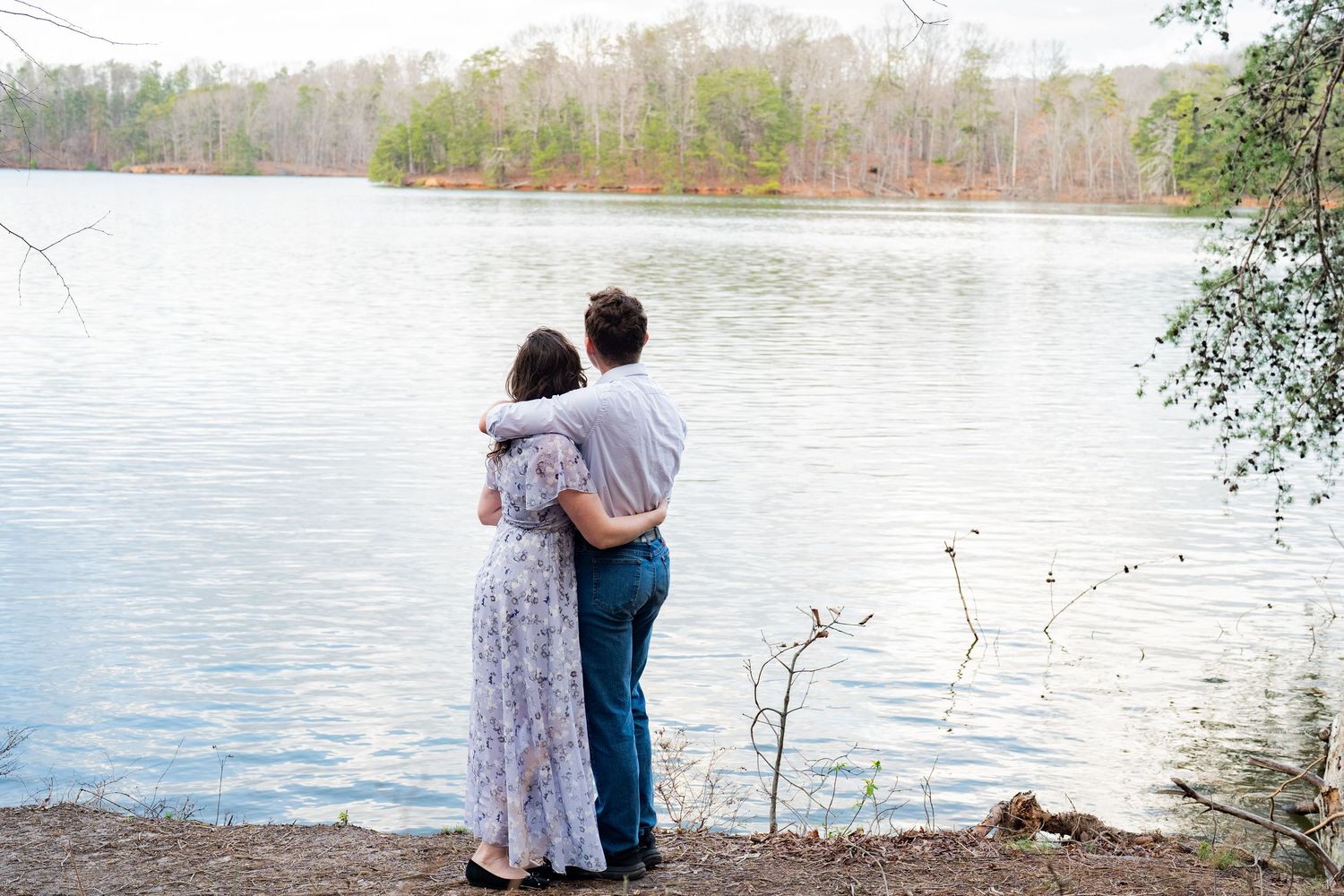 Two people embrace while looking out over a peaceful lake surrounded by trees on a calm day.