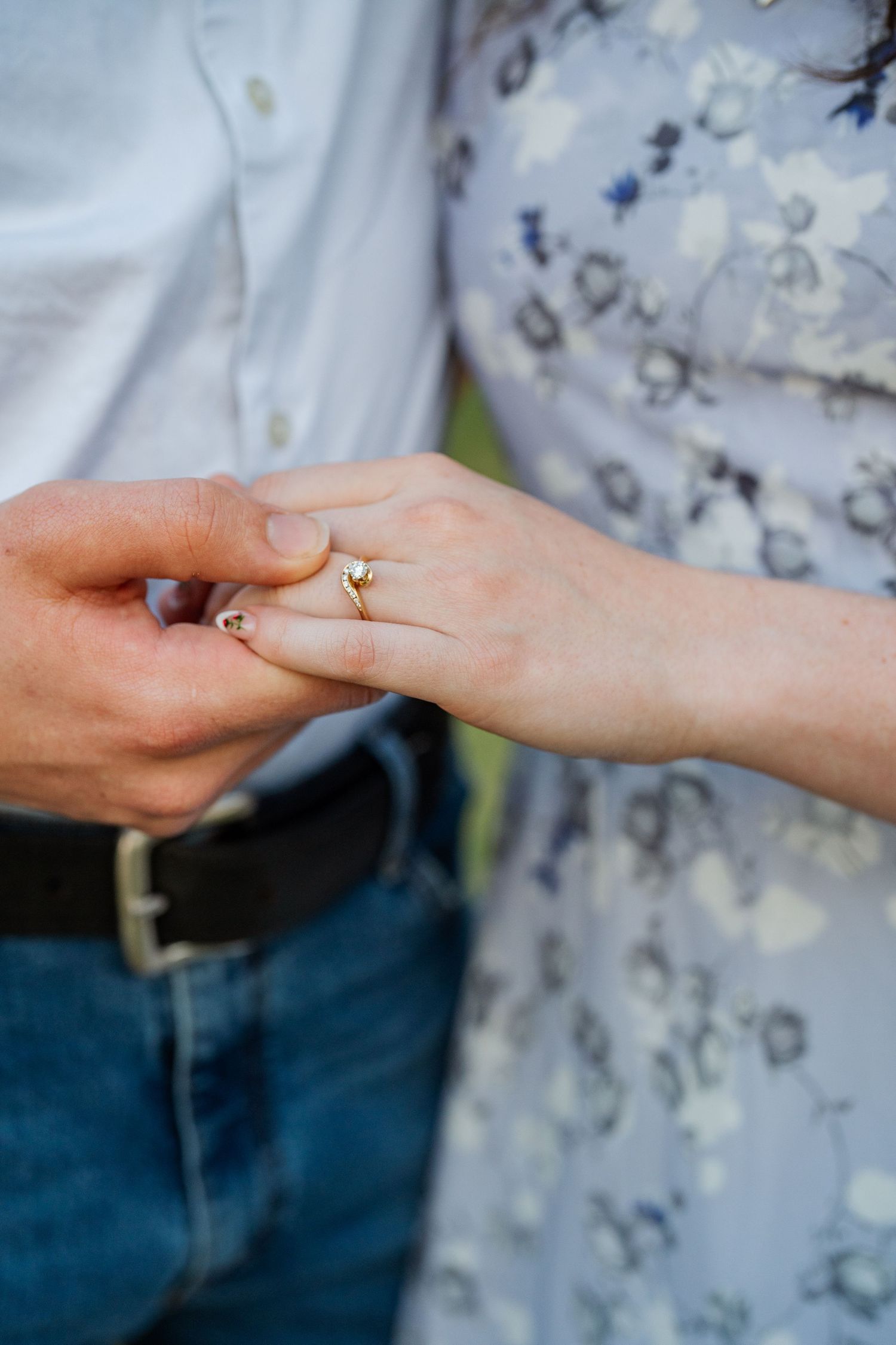 Close-up of hands exchanging an engagement ring, with a floral dress and denim visible in the background.