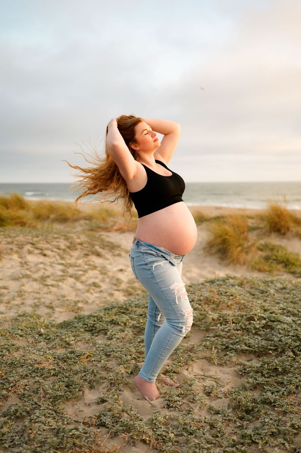woman poses during maternity photoshoot on top of Ventura beach dunes in sports bra and jeans showing her bare belly, wind blowing her hair