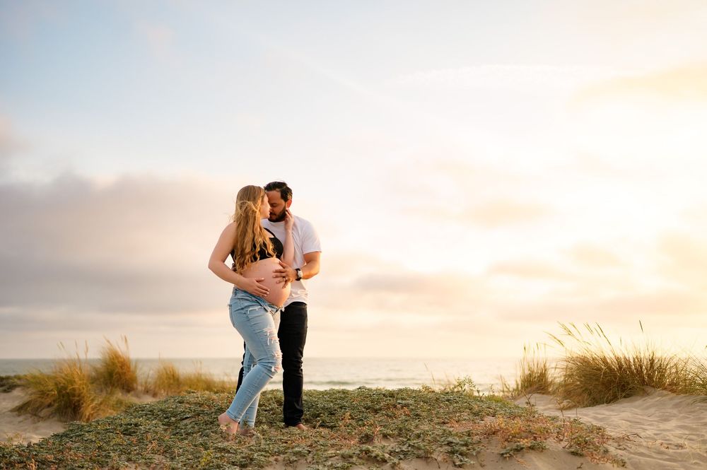 couple embraces during maternity photoshoot on top of Ventura beach dunes. woman wears jeans and sports bra showing her bare belly
