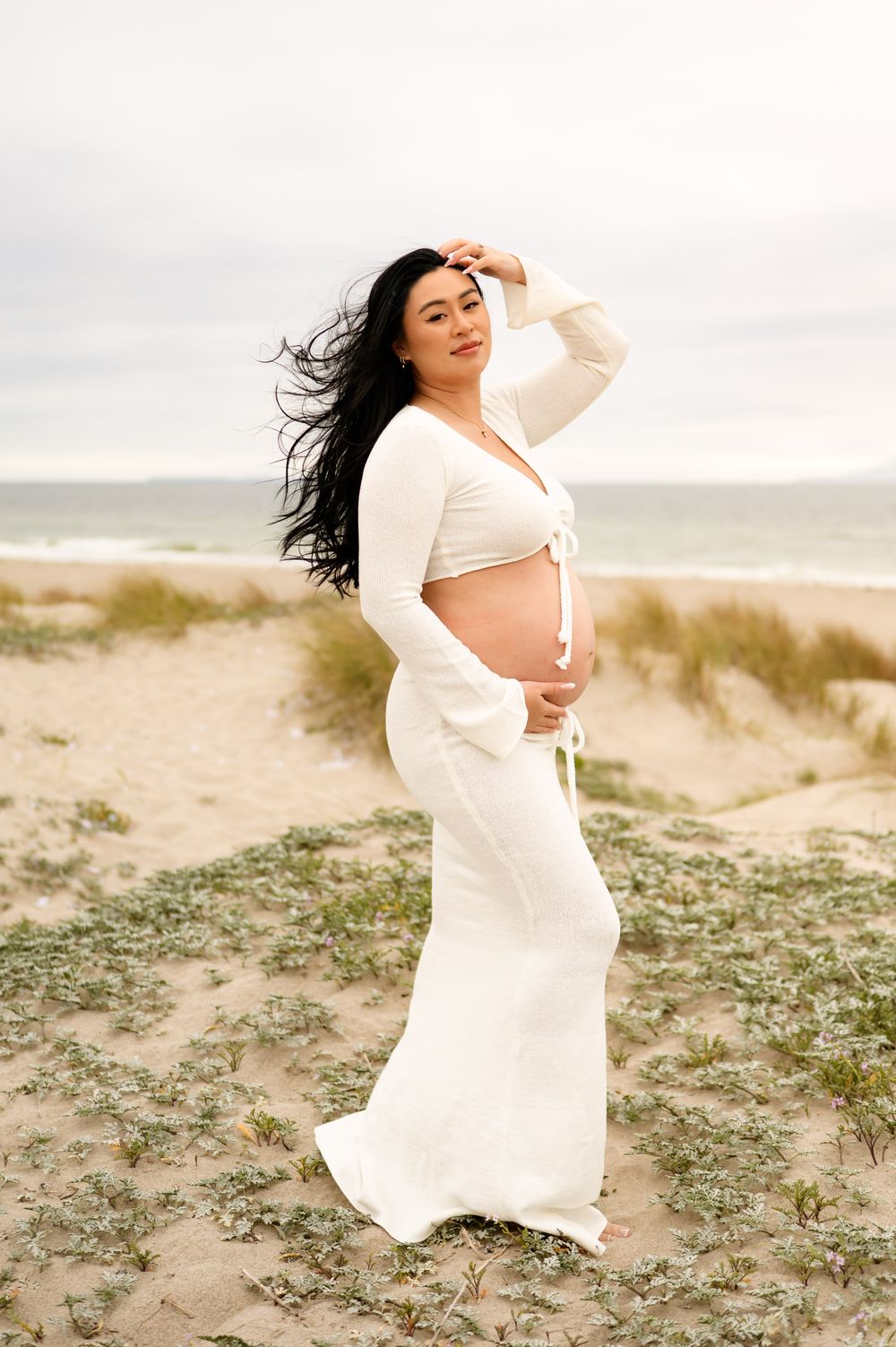 Woman in jluxe crochet dress is posed on sand dunes during her maternity photoshoot while on her Santa Barbara babymoon