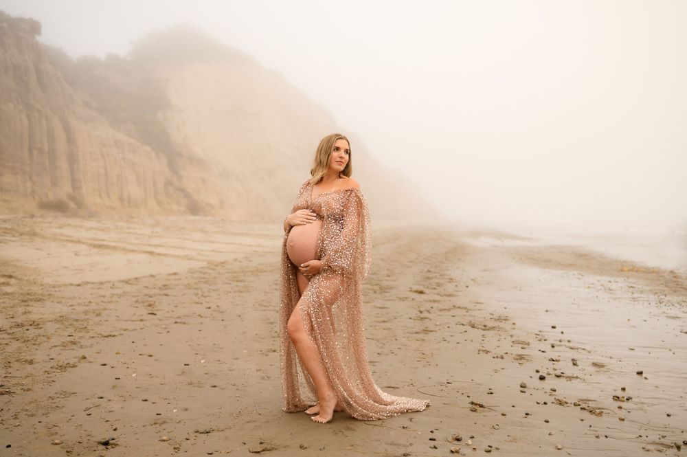 Carpinteria maternity photoshoot by Ventura Photographer of woman posed in sparkle dress at Loon Beach in the fog holding bare belly