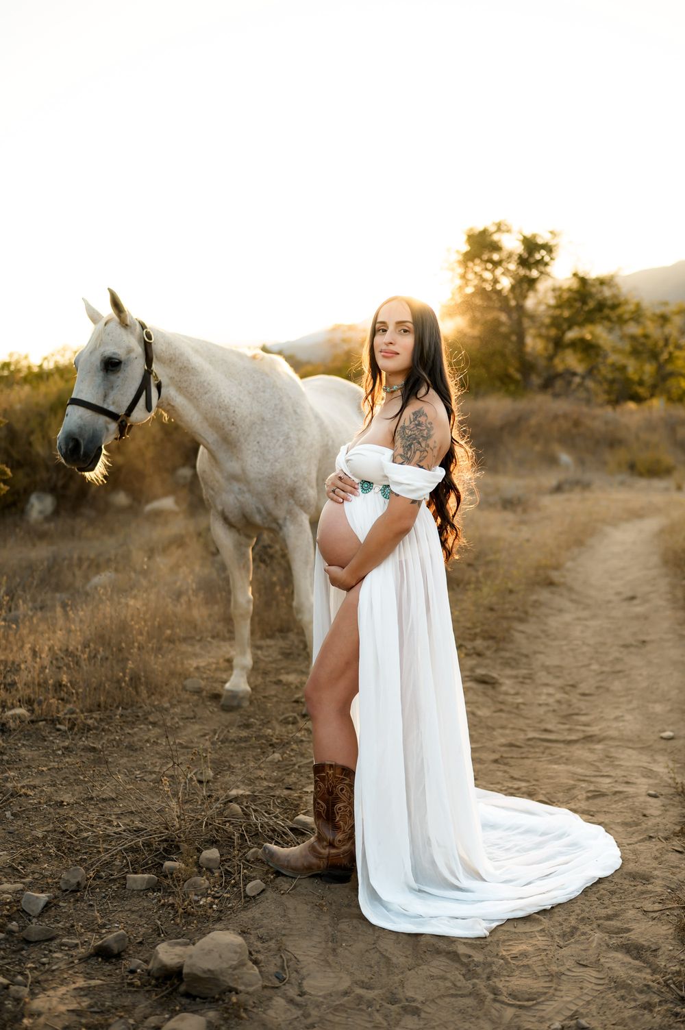 Ojai river preserve maternity photoshoot of woman in white dress and cowboy boots holding her belly in front of a white horse during sunset