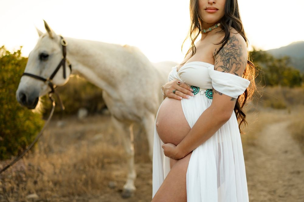 Ojai river preserve maternity photoshoot of woman in white dress holding her belly in front of a white horse during sunset