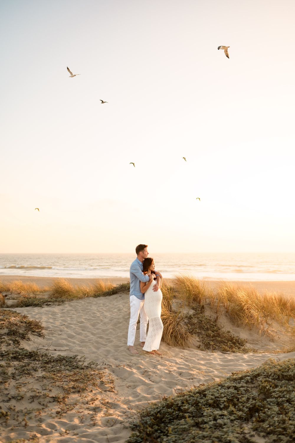 Ventura beach maternity photoshoot of couple posed on Oxnard beach dunes holding belly at sunset with birds flying around them