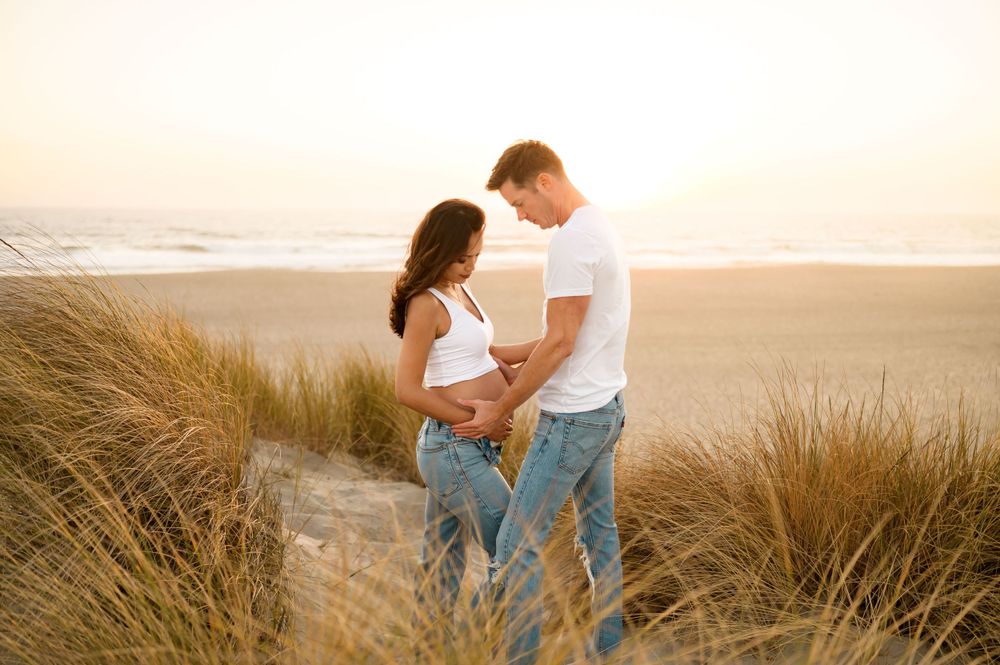 Ventura beach maternity photoshoot of couple in white t-shirts and jeans posed on Oxnard beach dunes holding belly at sunset