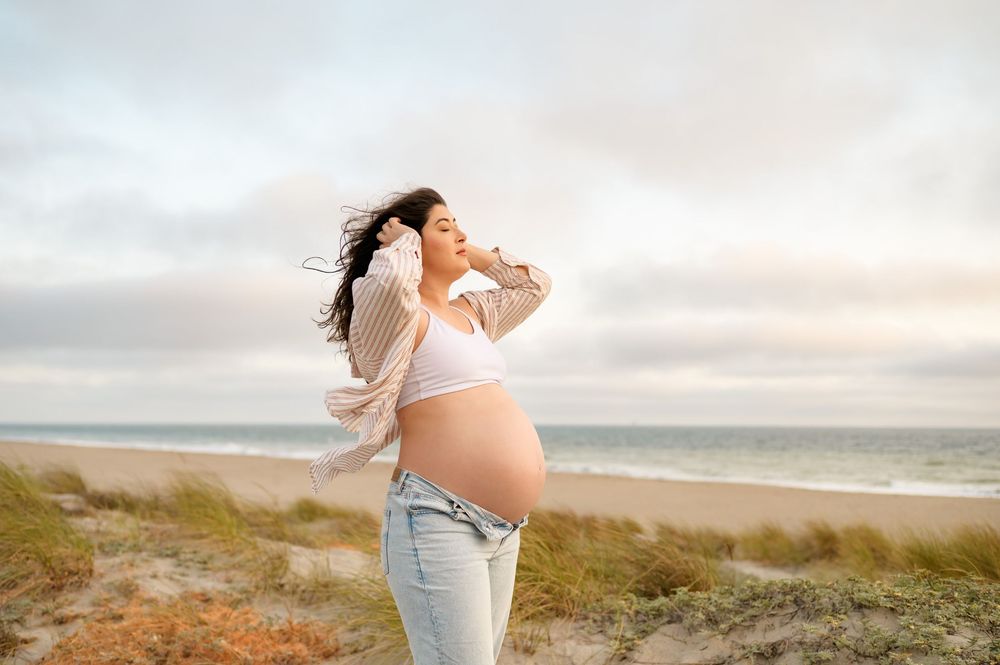 woman poses in wind during maternity photoshoot on top of Carpinteria beach dunes. woman wears jeans and sports bra showing her bare belly
