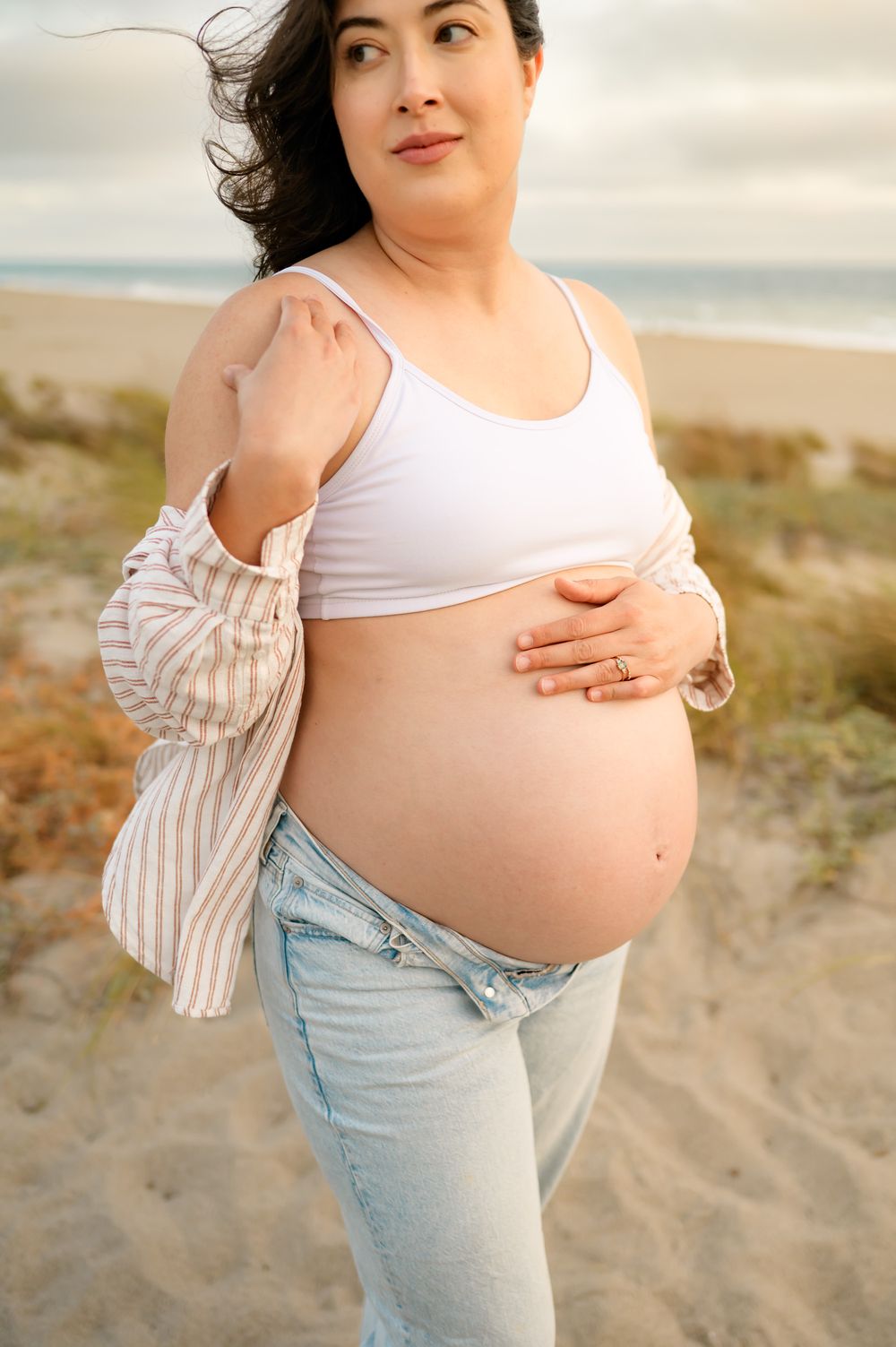 woman poses in wind during maternity photoshoot at Ventura beach. woman wears jeans and sports bra showing her bare belly