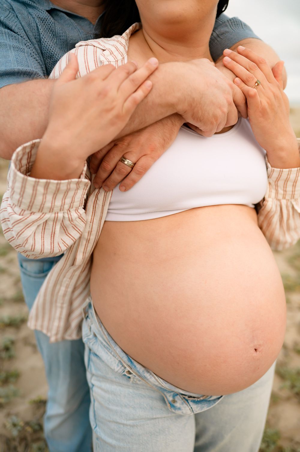 couple embraces during maternity photoshoot on top of Ventura beach dunes. woman wears jeans and sports bra showing her bare belly