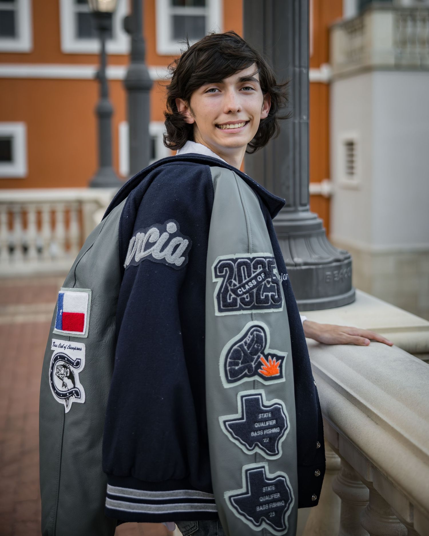 Portrait of someone in a burgundy and gray varsity jacket leaning against a brick wall with bokeh lights in background.