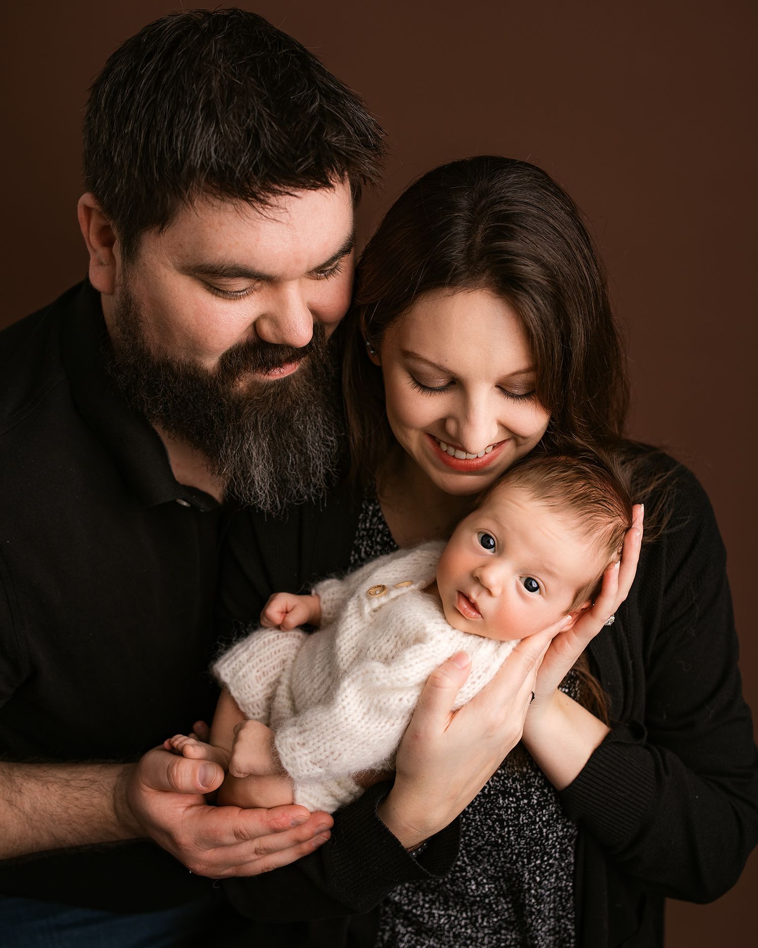 A bearded father and dark-haired mother lovingly gaze at their newborn baby dressed in white knit outfit.