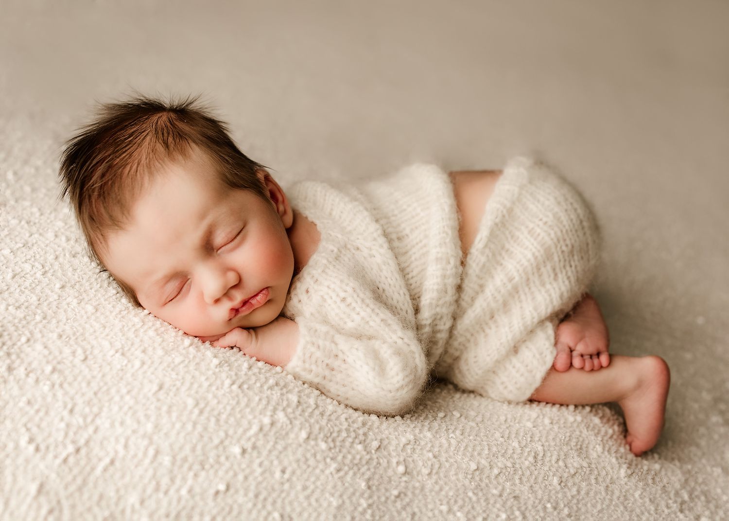 Peaceful newborn baby sleeping curled up in cream knit romper on soft white blanket.
