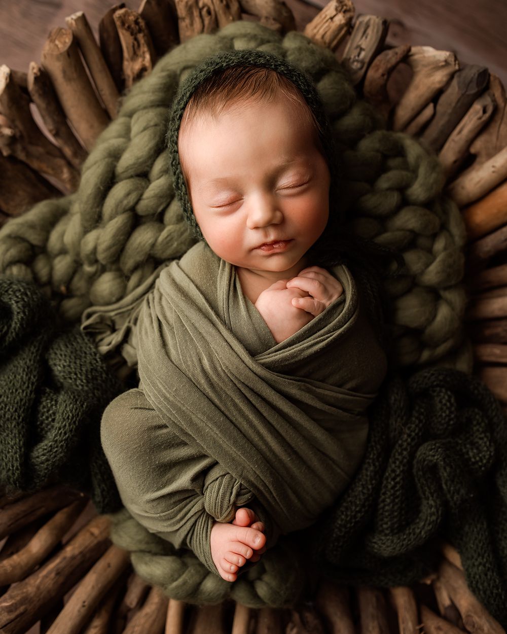 Newborn baby swaddled in olive green wrap sleeping peacefully in a rustic wooden bowl with chunky knit blanket.